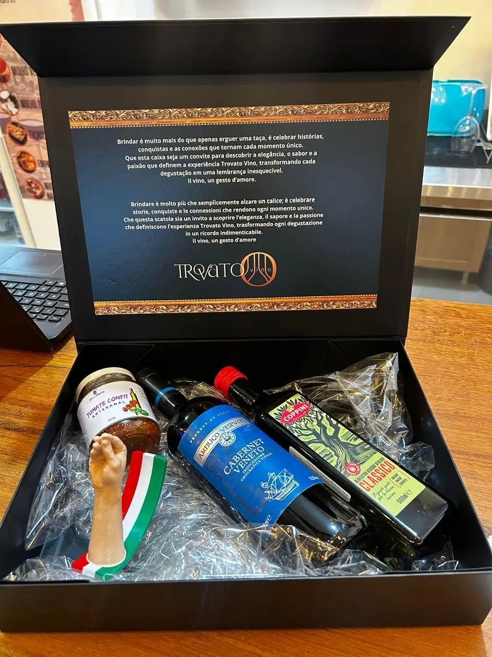 Caixa preta aberta com a mensagem da Trovato Vino na tampa, um Cabernet Veneto, azeite extravirgem, tomate confit e um enfeite com as cores da Itália