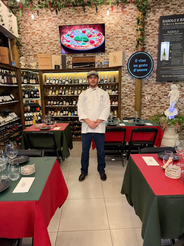 Júlio de dólmã branco no salão da Trovato Vino, entre mesas postas de vermelho e verde e estantes de vinho
